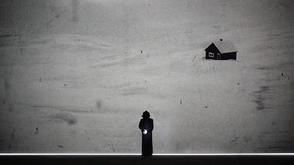 Ein Mann in schwarzem Anzug und Hut schaut zu einer riesigen Leinwand auf, auf der ein einsames Haus im Schnee zu sehen ist.