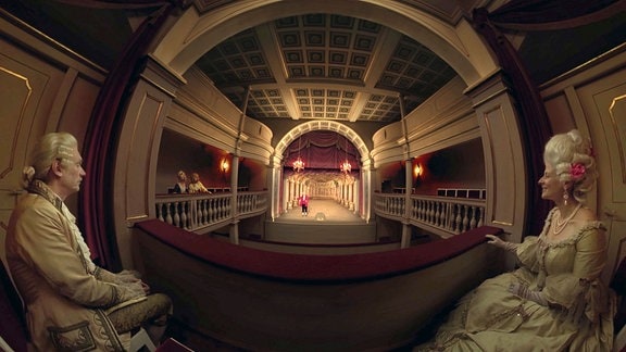 Blick in einen Saal mit Bühne. Links und rechts sitzen Personen in historischen Kostümen.