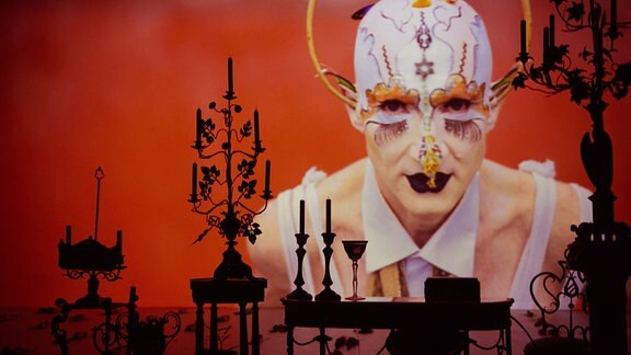 Gesicht einer hell geschminkten Person mit weißer, aufwändig dekorierter Maske auf einer roten Leinwand. Davor auf einer Bühne die Umrisse von barock anmutenden Möbeln.