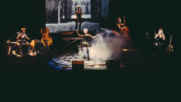 Auf einer dunklen Theaterbühne performen fünf Musikerinnen mit weiß geschminkten Gesichtern.