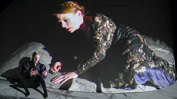 Eine übergroße Frau im Brokat-Kleid beugt sich in einer Videoprojektion zu einem hockenden Mann auf der Bühne.