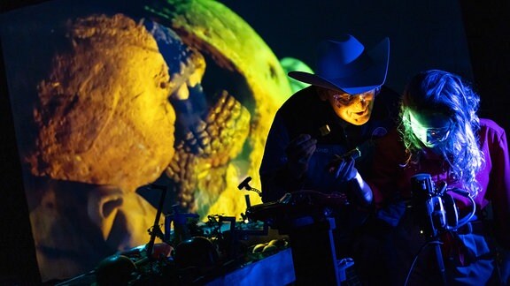 Ein Mensch mit einem breitkrempigen Cowboy-Hut hantiert mit einer Kamera, die ein grün-gelbes Bild im Hintergrund projiziert.