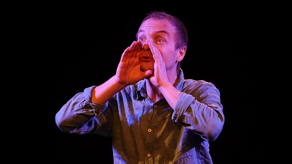 Zu sehen ist ein Mann in blauem Hemd, der beide Hände zu einem Trichter am Mund formt; er wird von Bühnenlicht beleuchet.