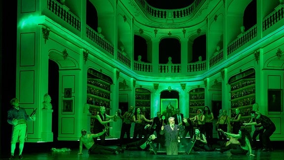 Ein grün beleuchteter Bibliothekssaal, in der Mitte versammelt sich eine Gruppe von Menschen und singt