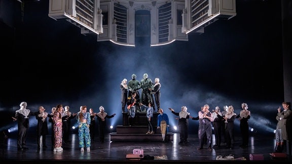 Menschen stehen auf einer Theater-Bühne links und rects neben einem Denkmal mit Goethe und Schiller, darüber schwebt ein Modell der Anna Amalia Bibliothek im dunklen Bühnen-Himmel.