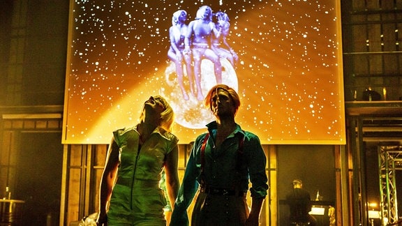 Zwei Menschen stehen Hand in Hand vor einer goldgelb erleuchteten Bühne und schauen nach oben. 