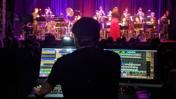 Ein Lichttechniker steuert am Pult die Beleuchtung für ein Konzert, die Bühne ist pink- und lilafarben beleuchtet