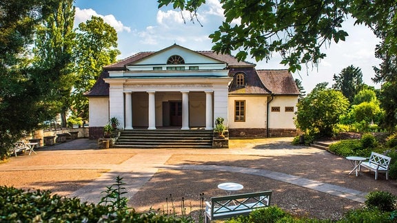 Ein historisches Gebäude mit 4 Runden Säulen im Eingang, umgeben von einem Park mit Sträuchern und Bänken