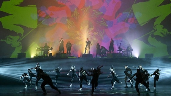  Schauspieler tanzen, im Hintergrund ist eine Band mit Macbeth zu sehen.  