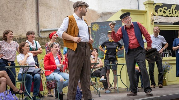 Szene aus dem Stück "Unter Nossen – Eine Stadt im Blaurausch": zwei Männer mit Westen und Mützen stehen auf einer Bühne und sind zum Publikum gerichtet