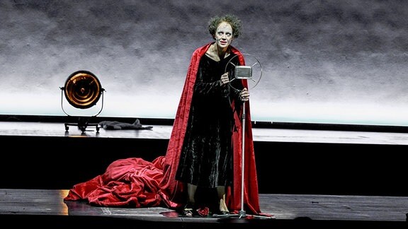 Eine Frau in schwarzem Kleid und mit rotem Umhang steht vor einem altertümlichen Mikrofon. Ihr Gesicht ist weiß geschminkt, ihre Haare sind grün gefärbt.