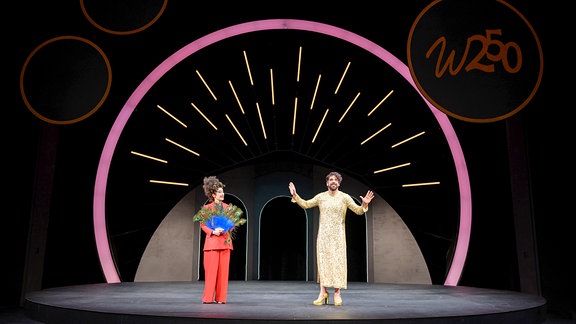Zwei Personen auf einer Bühne. Als Bühnenbild ist ein leuchtender rosa Kreis zu sehen.