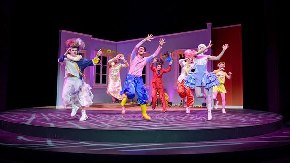 Menschen in bunter Kleidung tanzen auf einer Bühne, im Hintergrund sind rosa Kulissen zu sehen.