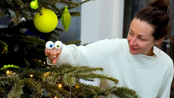 Eine Frau steht an einem Weihnachtsbaum und formt mit ihrer Hand ein Klappmaul mit zwei Plastik-Augen.
