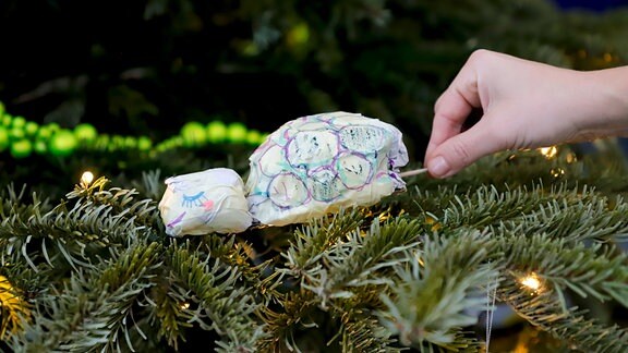 Eine angemalte Schildkrötenpuppe aus Papier wird von einer Hand auf einem Weihnachtsbaum drapiert.
