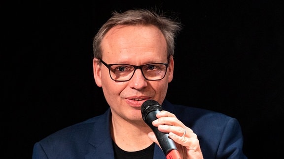 Ein Mann mit Brille steht vor einem schwarzen Hintergrund mit einem Mikrophon in der Hand und lächelt.  