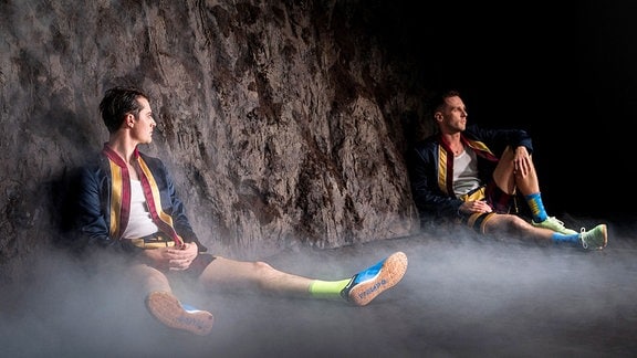 Zwei Fußballspieler lehnen im Sitzen umgeben von Nebel an einem braunen Berg.