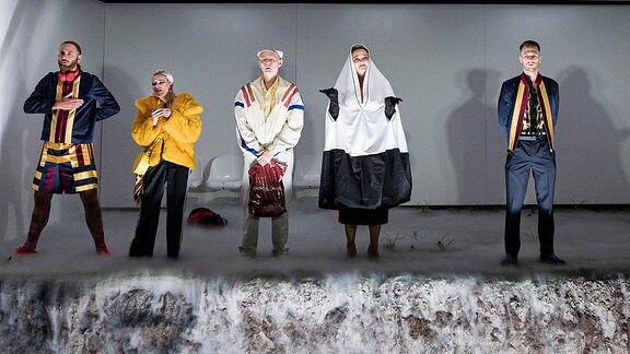Sechs Personen stehen in unterschiedlichen Kostümen auf einer Theaterbühne nebeneinander, vor ihnen eröffnet sich ein Abgrund.