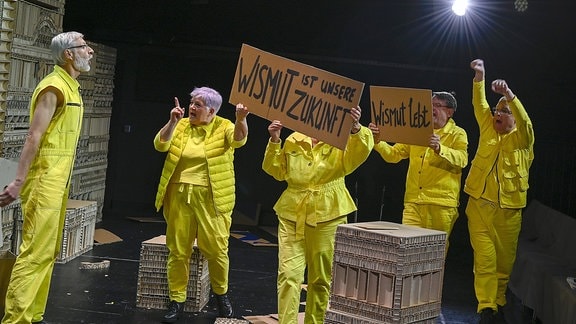 In "Tausend Sonnen" an der Bürgerbühne Dresden stehen mehrere Menschen in gelben Anzügen auf der Theaterbühne und halten Schilder hoch mit Aufschriften wie "Wismut lebt".