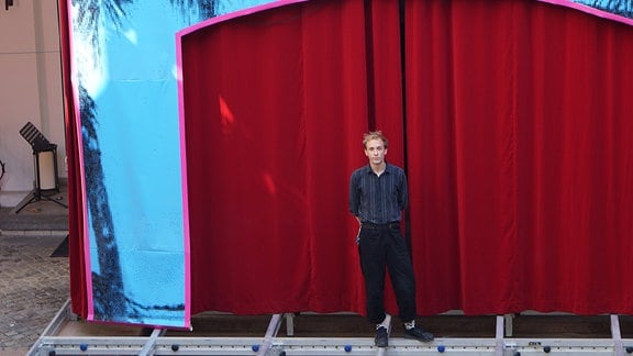 Ein junger blonder Mann steht auf einem Gerüst vor einem roten Theatervorhang
