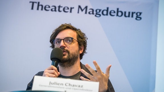Julien Chavaz spricht in ein Mikrofon und gestikuliert, hinter ihm steht groß "Theater Magdeburg".
