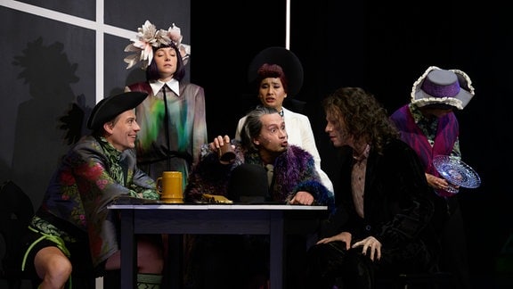 Szene aus einem Theaterstück: Mehrere Menschen sitzen an einem runden Tisch zusammen und reden angeregt.