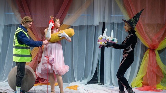 Eine als Hexe verkleidete Frau läuft mit einer Torte über eine Bühne, auf der eine Frau im rosa Tüllkleid und ein Mann in gelber Warnweste stehen.