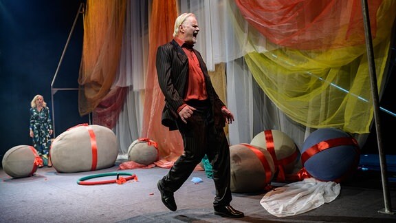 Ein Schauspieler in braunem Nadelstreifenanzug und rotem Hemd geht schreiend über eine mit großen Stoffbällen und bunten Tüchern gestaltete Bühne
