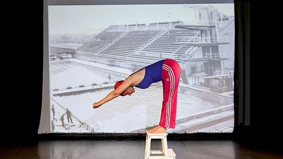 Das Bild zeigt die Künstlerin Lykke Langer mit einer roten Sporthose, einem blauen Badeanzug und einer Schwimmkappe. Sie deutet einen Kopfsprung an. 