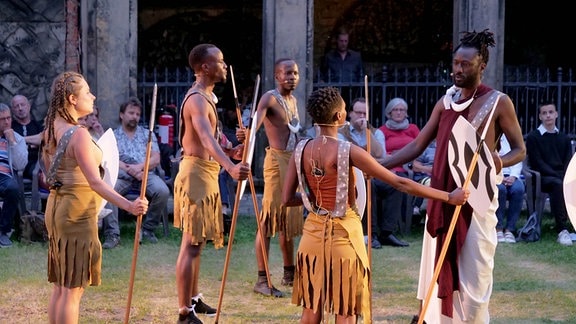 Eine Gruppe Schauspielerinnen und Schauspieler unterschiedlicher Hautfarbe mit afrikanischen Gewändern und Speeren steht vor Publikum in einem Kreis