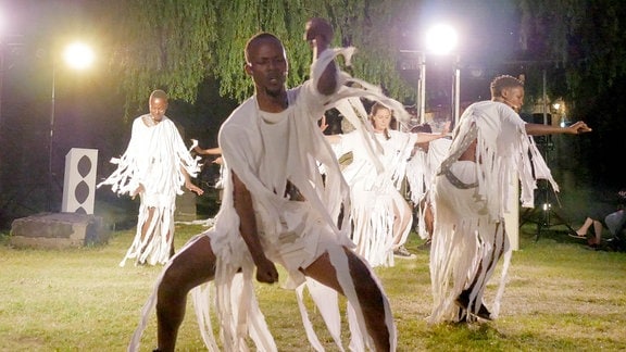 Eine Gruppe Schauspielerinnen und Schauspieler unterschiedlicher Hautfarbe in langen weißen Gewändern tanzt auf dem Rasen einer Freiluftbühne