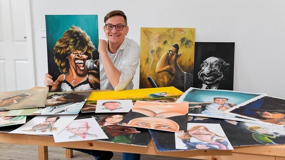 Thomas Leibe, sitzt an einem Tisch, umgeben von Karikaturen welche er gezeichnet hat.