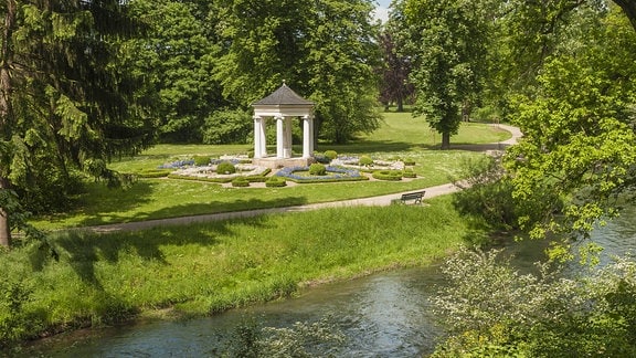 Zu sehen ist der Blick in den Schlosspark Tiefurt, im Hintergrund steht der Musentempel am Fluss, Bäume ragen ins Bild