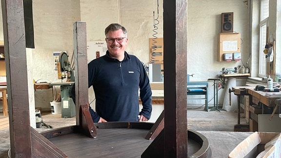 Zu sehen ist Udo Vogel, Tischlermeister und Chef der Bau- und Möbeltischlerei Thier im Leipziger Osten, wie er hinter einem umgedrehten Tisch in seiner Werkstatt steht.