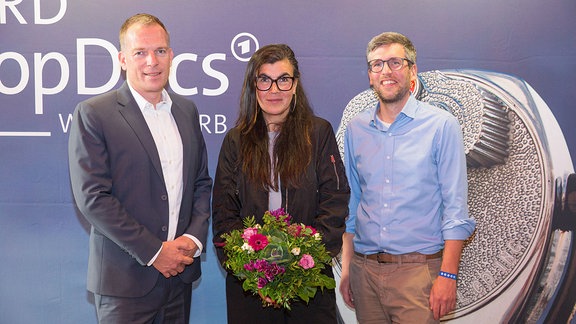 Drei Personen stehen vor einer Aufstellwand. Die Person in der Mitte hält einen Blumenstrauß.