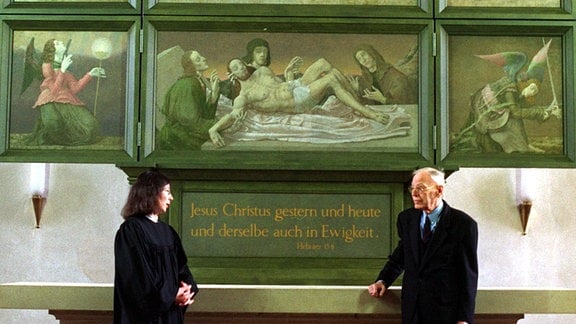 Eine Pfarrerin und ein Mann stehen in einer Kirche unter einem Altar mit aufgeklappten Flügeln, der biblische Szenen zeigt.