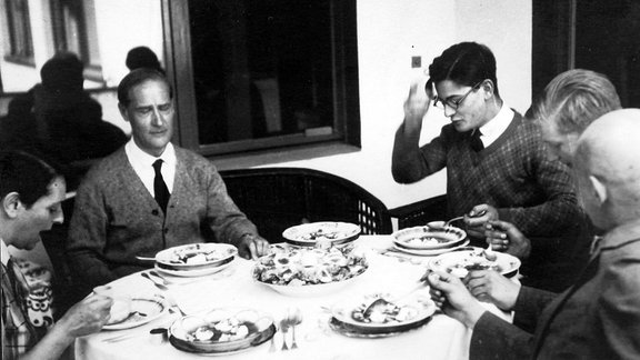 Familie Feininger sitzt an einem runden Tisch, Geschirr mit Essen steht darauf.