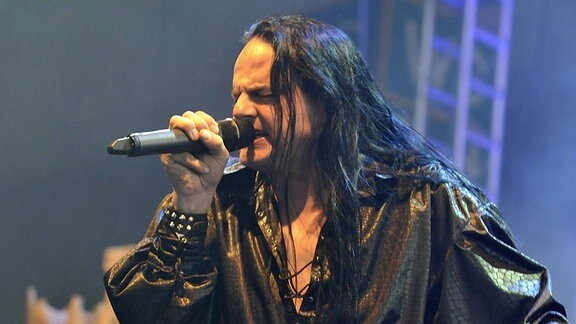 Ein Mann mit langem schwarzem Haar und schwarzer Kleidung singt in ein Mikrofon.
