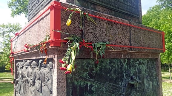 Am Sockel eines Denkmales hängen Blumen. 