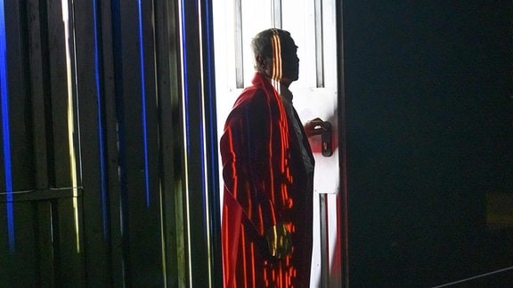 Ein Mann in einem roten Bademantel steht auf einer Bühne und öffnet eine Tür in einer schwarzen Wand.