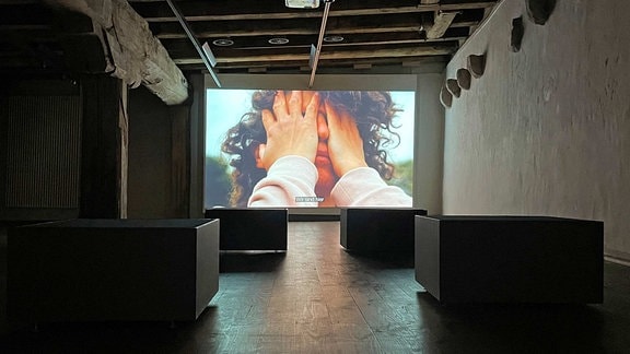 Man blickt auf eine Videoinstallation in der Galerie Waidspeicher, auf dem Bildschirm ist eine Frau zu sehen, die die Hände vor das Gesicht schlägt.