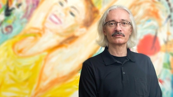 älterer Mann mit grauem Haar steht vor einem bunten Gemälde