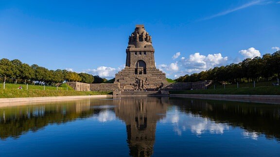 Das Völkerschlachtdenkmal in Leipzig, davor ein künstlich angelegter Teich, im Hintergrund blauer Himmel