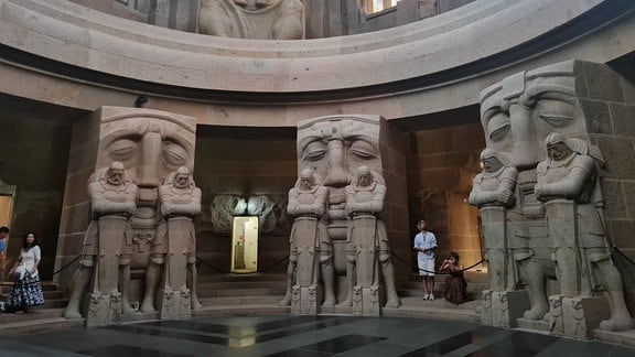 Völkerschlachtdenkal Leipzig im Inneren: eine runde Halle mit imposanten Figuren aus Sandstein. Sie haben die Arme verschränkt und blicken nach unten.