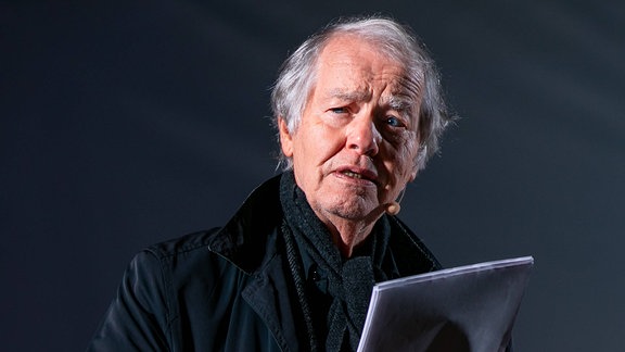 Ein Mann im Mantel und mit grauen Haaren hält mehrere Blätter Papier in seiner Hand.