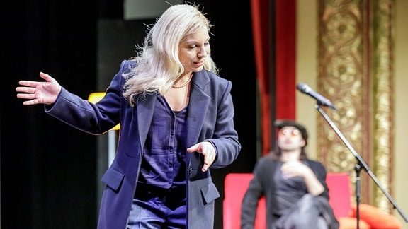 Eine Frau mit blonden Haaren und in einem langen blauen Mantel spricht gestikulierend.