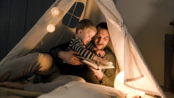 Vater liest seinem Sohn in einem beleuchteten Zelt zu Hause ein Buch vor.