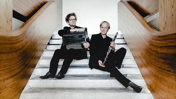 Klarinettist Uroš Rojko (rechts) und Akkordeonist Luka Juhart (links) sitzen auf einer weißen Treppe. Sie halten ihre Instrumente in der Hand. Beide sind schwarz gekleidet. 