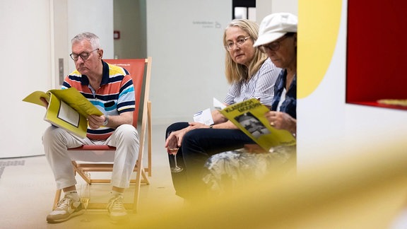 Ein Mann sitzt in einem Liegestuhl und blättert in einer Zeitschrift, rechts neben ihm sitzen zwei Frauen