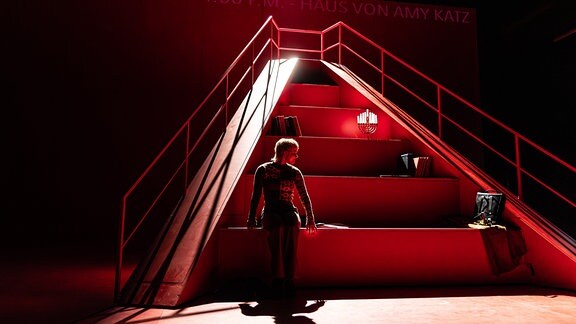 Eine Person steht am Fuße einer in rotes Licht getauchten Treppe in Form einer Bugspitze.